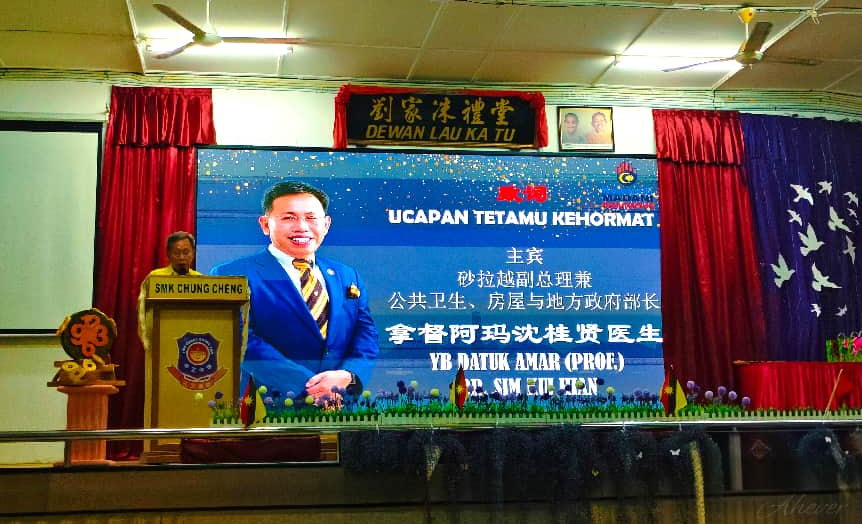 身穿黄色服装的发言人在礼堂讲台致辞，后方屏幕展示活动嘉宾信息。
