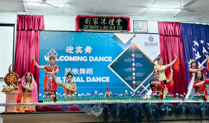 身穿传统表演服装的舞者在礼堂舞台表演，背景屏幕写有迎宾舞。
