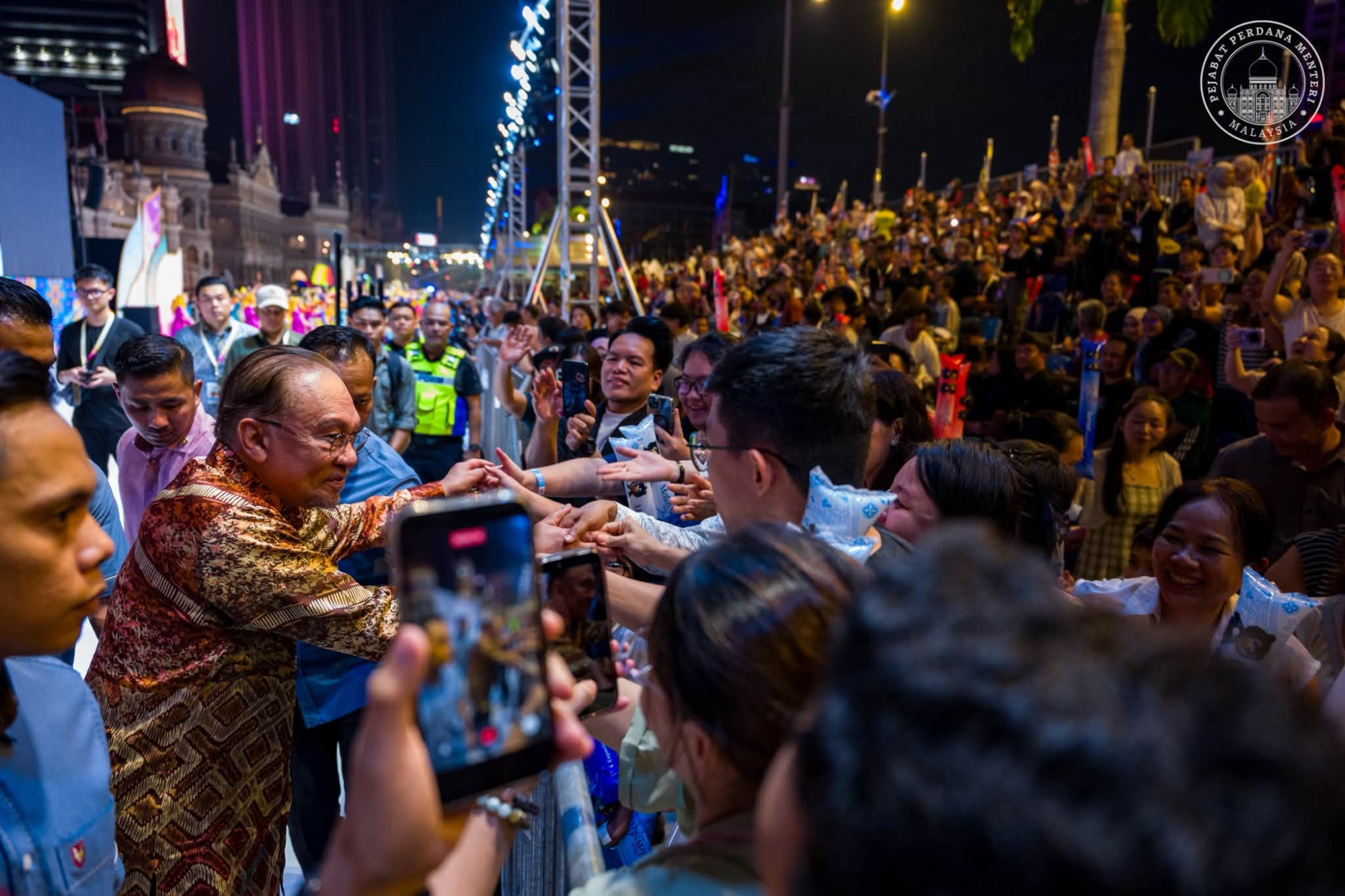 夜间活动现场，穿花纹衬衫的嘉宾与围栏旁观众互动，观众举起手机拍照。